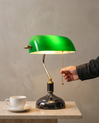 Green Banker desk lamp, emerald green glass shade, brass arm, dark chrome base — in use, hand on the pull-chain | Dalfa CH/FR