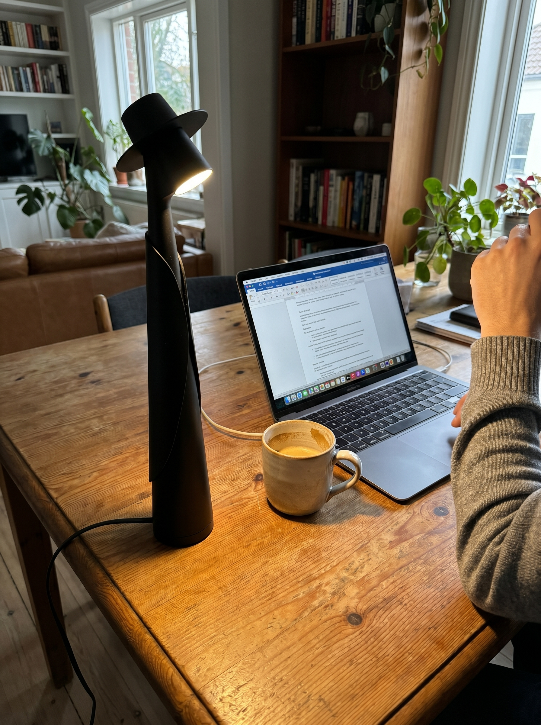 Matte black figure lamp lit on a wooden home-office desk beside a laptop and mug — Dalfa CH/FR