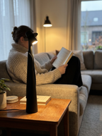 Matte black figure lamp lit on a side table while a person reads on the sofa, evening — Dalfa CH/FR