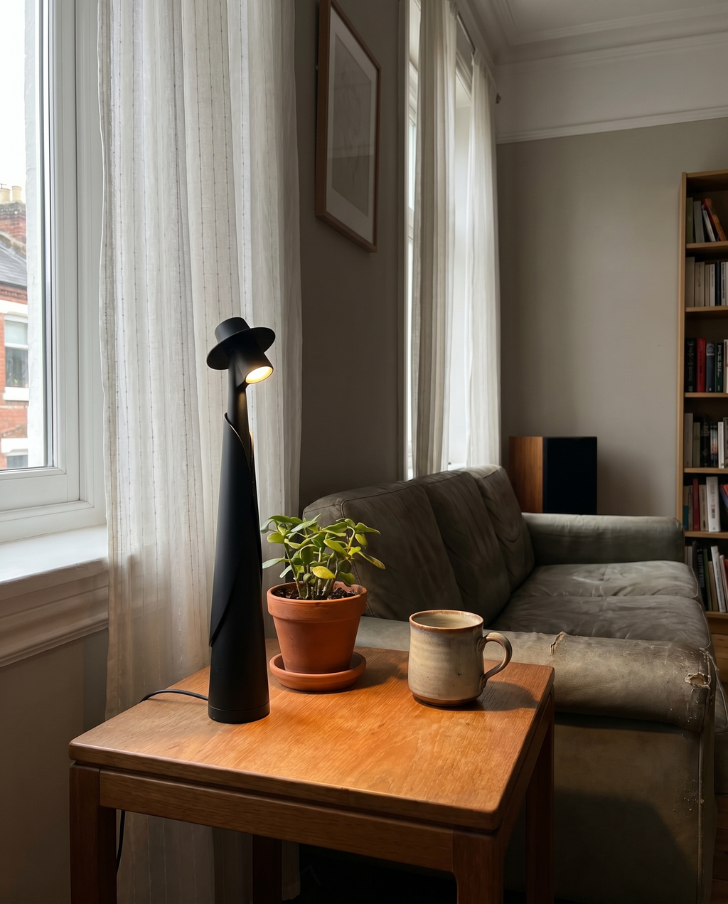 Matte black figure lamp lit on a side table by a window with a plant, real living room — Dalfa CH/FR