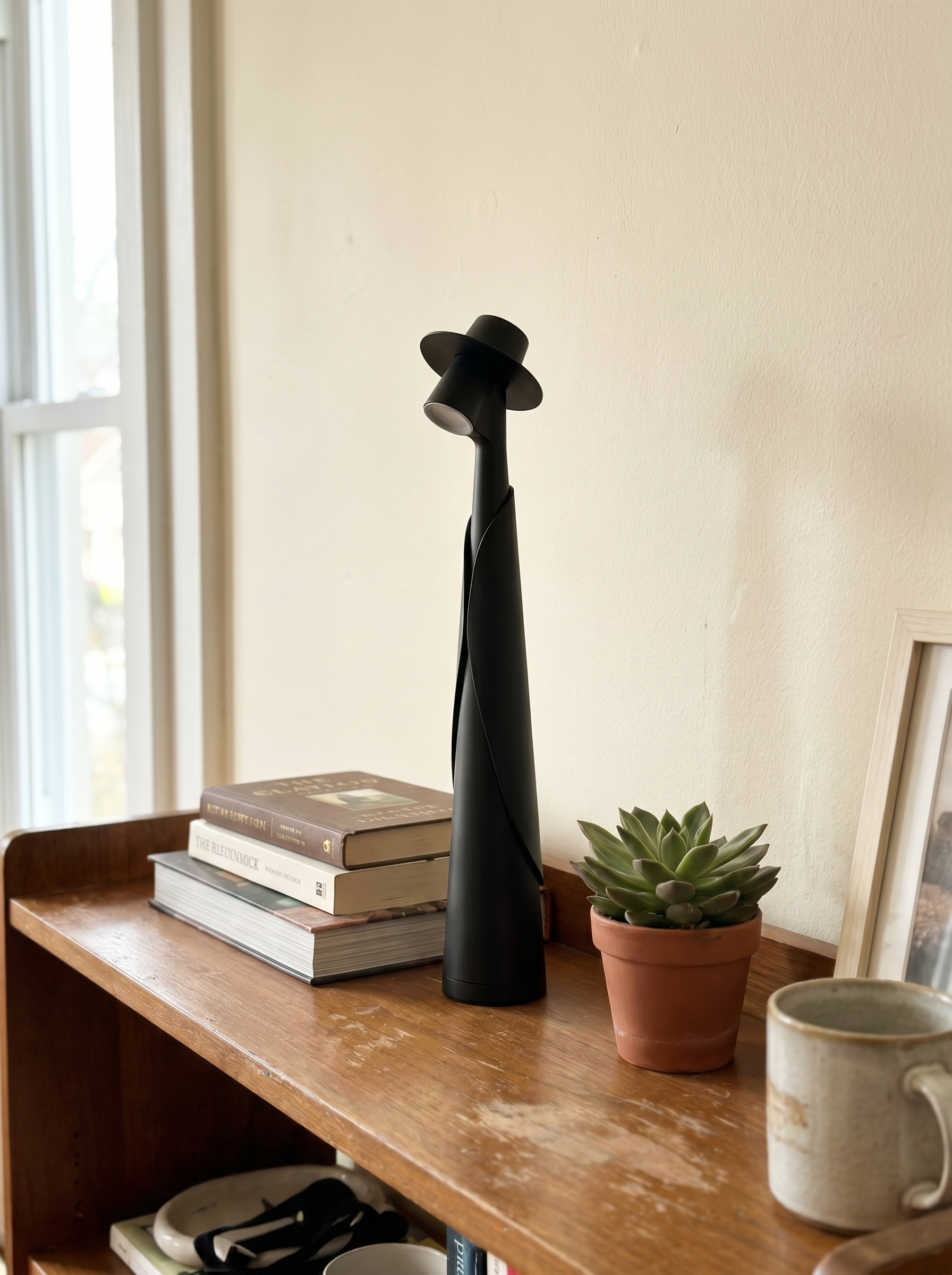 Matte black figure lamp off on a shelf with books and a plant, real home — Dalfa CH/FR